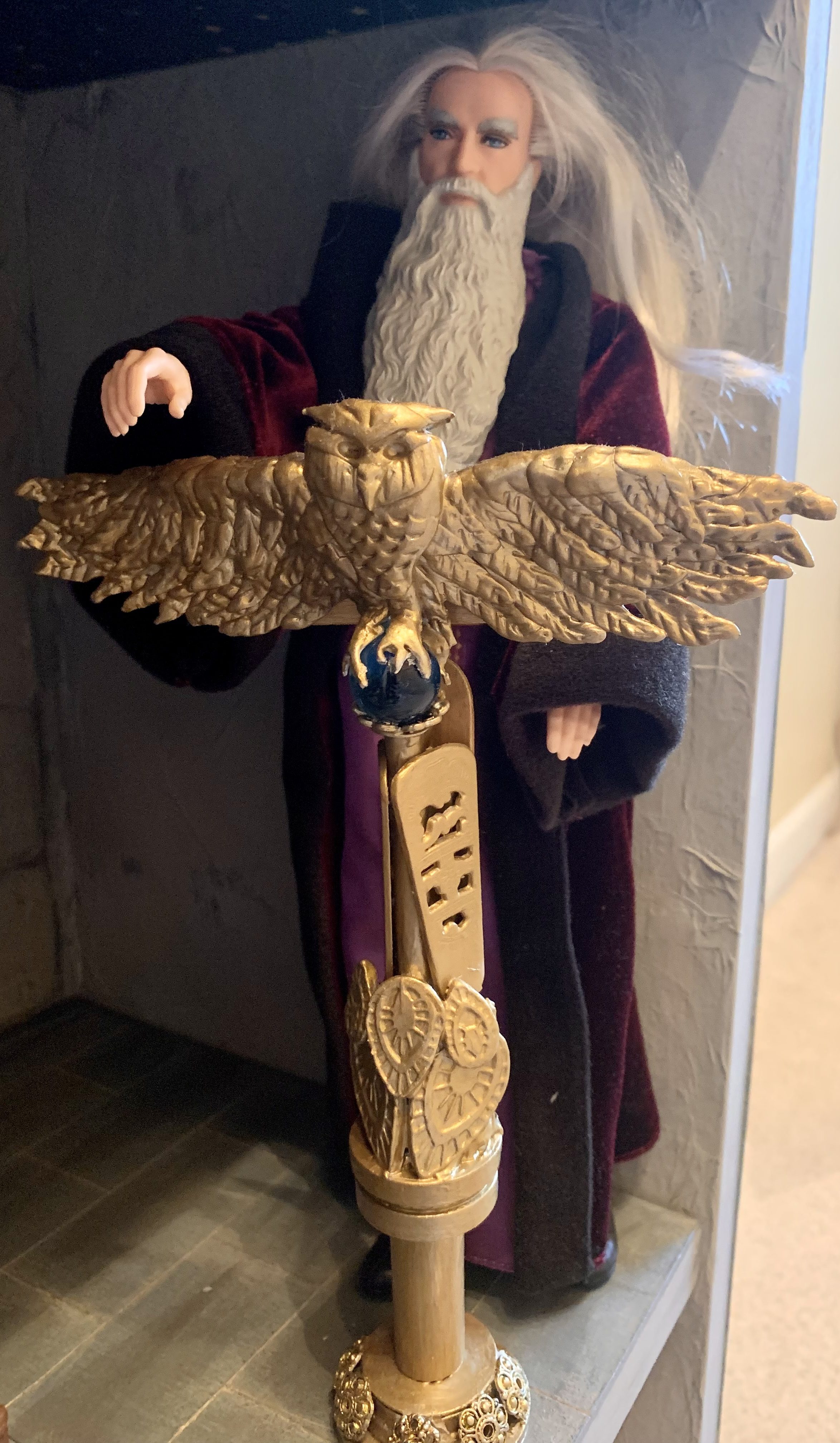 A Dumbledore doll standing at his podium, made from polymer clay and various other items. The top portion of the podium features an owl with its wings spread out, sitting on a blue orb. 