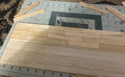 Trimmed popsicle sticks being glued onto a thin balsa wood base, in the style of a wood floor. 
