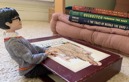 A Harry Potter doll sitting at a make-shift table made from the sleeve of an American Girl book set.
