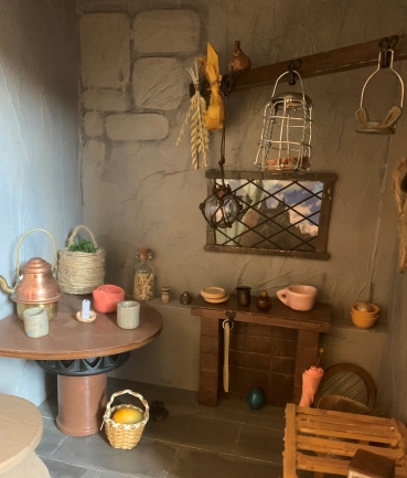 A view of a corner of a miniature Hagrid's Hut dollhouse room. A table sits against the wall, and has a candle, copper tea kettle, and cups on it. 