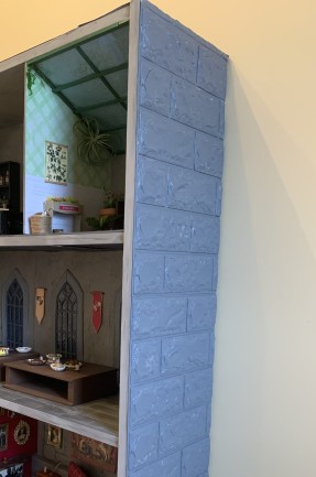 Portion of a castle dollhouse made from a bookcase, with an outer wall showing adhesive faux bricks painted gray. 
